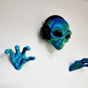 May include: A green and blue alien head wearing black headphones, with two matching green and blue hands on either side.