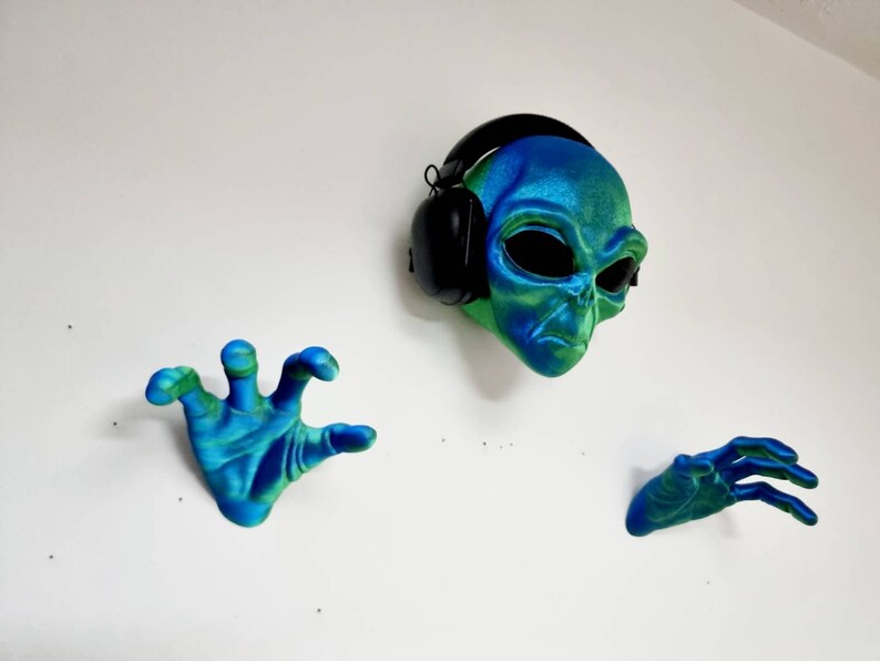 May include: A green and blue alien head wearing black headphones, with two matching green and blue hands on either side.