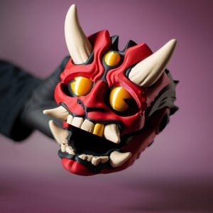 May include: A red and black Oni mask with white horns and teeth. The mask has three yellow eyes and a wide open mouth. The mask is held by a gloved hand.