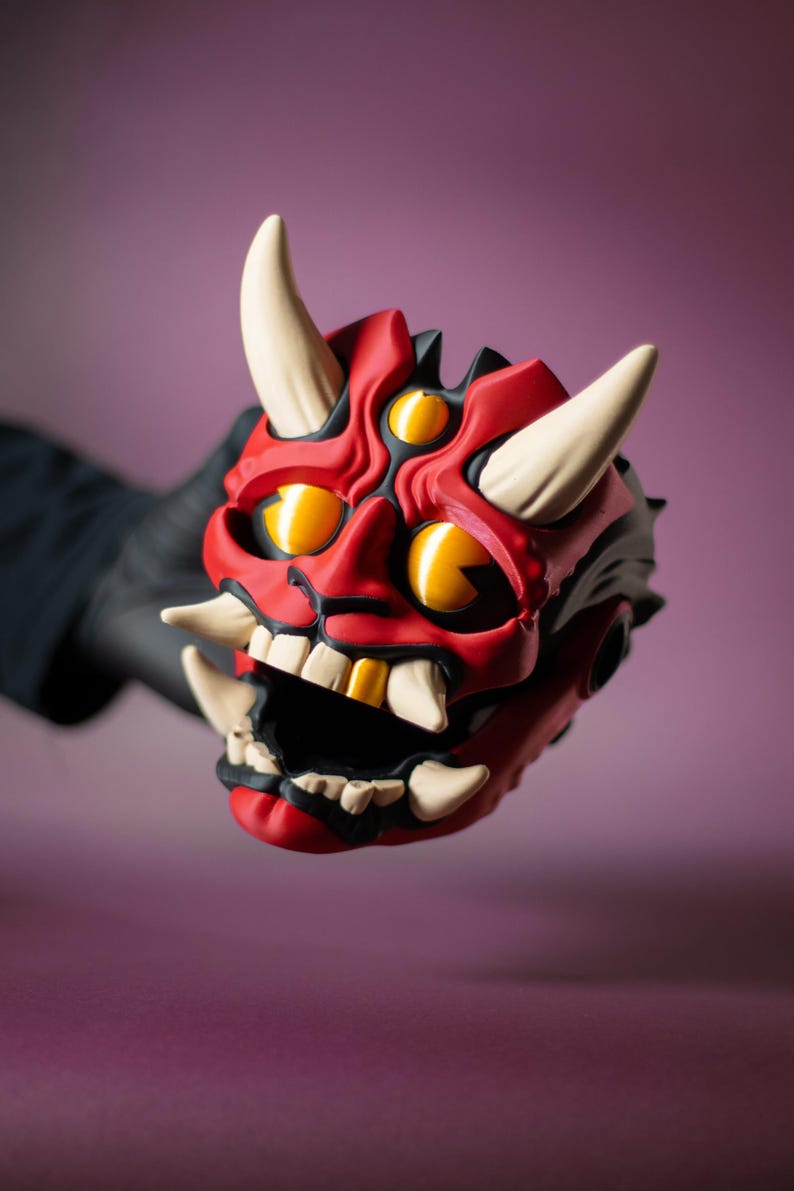 May include: A red and black Oni mask with white horns and teeth. The mask has three yellow eyes and a wide open mouth. The mask is held by a gloved hand.