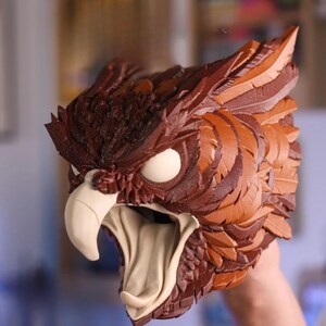 May include: A detailed, brown and tan owl mask with a fierce expression. The mask features textured feathers, large white eyes, and a prominent beak. The mouth is open, revealing a tan interior, creating a striking, realistic appearance. This mask is ideal for cosplay or costume events.