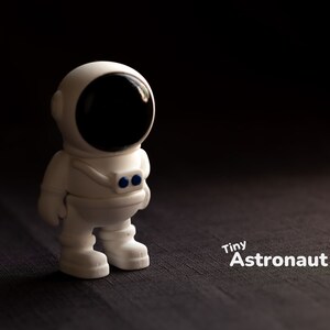 May include: A white plastic toy astronaut figurine with a black visor and blue details. The figurine is standing on a dark brown textured surface. The text "Tiny Astronaut" is written in white below the figurine.