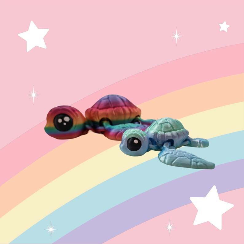 Puede incluir: Dos peque&ntilde;as figuras de tortugas de resina de colores. La tortuga m&aacute;s grande es de color arco&iacute;ris y la m&aacute;s peque&ntilde;a es azul y verde. Las tortugas est&aacute;n sobre un fondo de arco&iacute;ris.