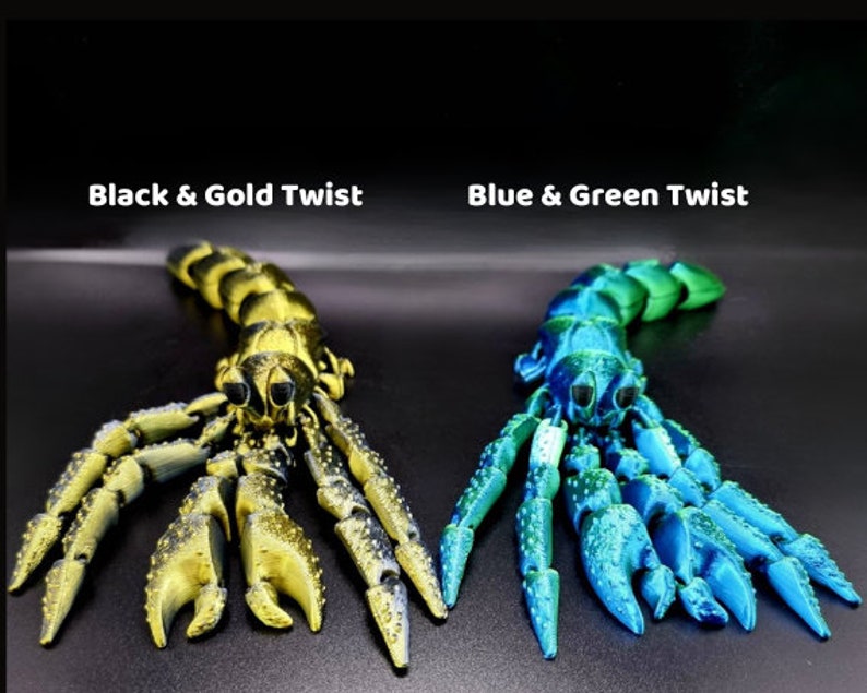 Puede incluir: Dos figuritas de escorpi&oacute;n impresas en 3D, una en negro y oro, la otra en azul y verde. Los escorpiones est&aacute;n en pose con sus garras extendidas.