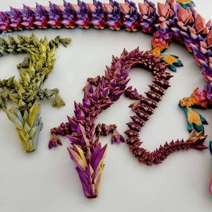 May include: Three origami dragons made of paper, each with a different color scheme. The dragons are arranged in a circle, with their heads pointing towards the center. The dragon on the left is green, the dragon in the center is purple, and the dragon on the right is red.