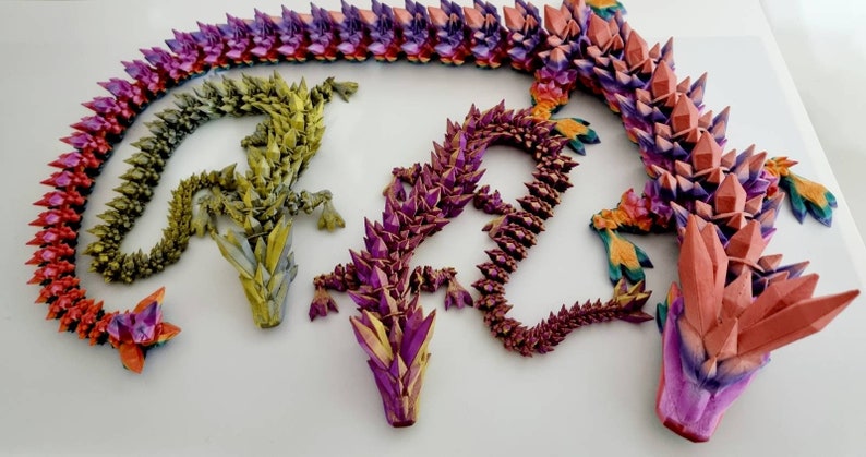 May include: Three origami dragons made of paper, each with a different color scheme. The dragons are arranged in a circle, with their heads pointing towards the center. The dragon on the left is green, the dragon in the center is purple, and the dragon on the right is red.