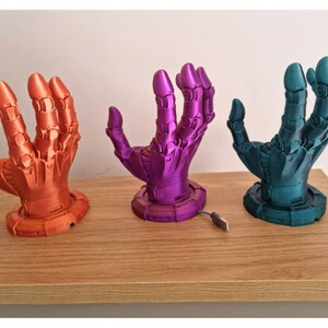 May include: Three 3D printed robot hands in different colors: orange, purple, and teal. The hands are posed with fingers extended and are mounted on a circular base. A USB cable is visible in the foreground.