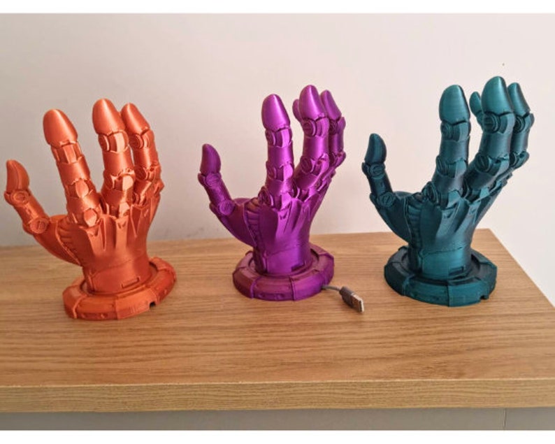 May include: Three 3D printed robot hands in different colors: orange, purple, and teal. The hands are posed with fingers extended and are mounted on a circular base. A USB cable is visible in the foreground.