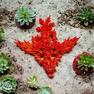 May include: A red and orange 3D printed dragon sculpture with a textured surface. The dragon is lying on a light brown surface with green plants around it.