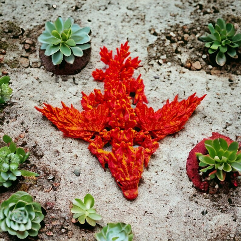 May include: A red and orange 3D printed dragon sculpture with a textured surface. The dragon is lying on a light brown surface with green plants around it.