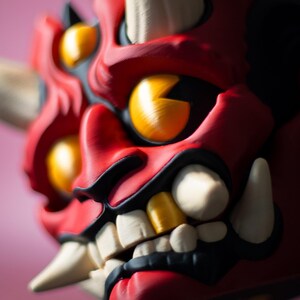 May include: A close-up of a red, black, and white Oni mask with yellow eyes and teeth. The mask has a fierce expression with sharp teeth and horns. The background is a soft pink.