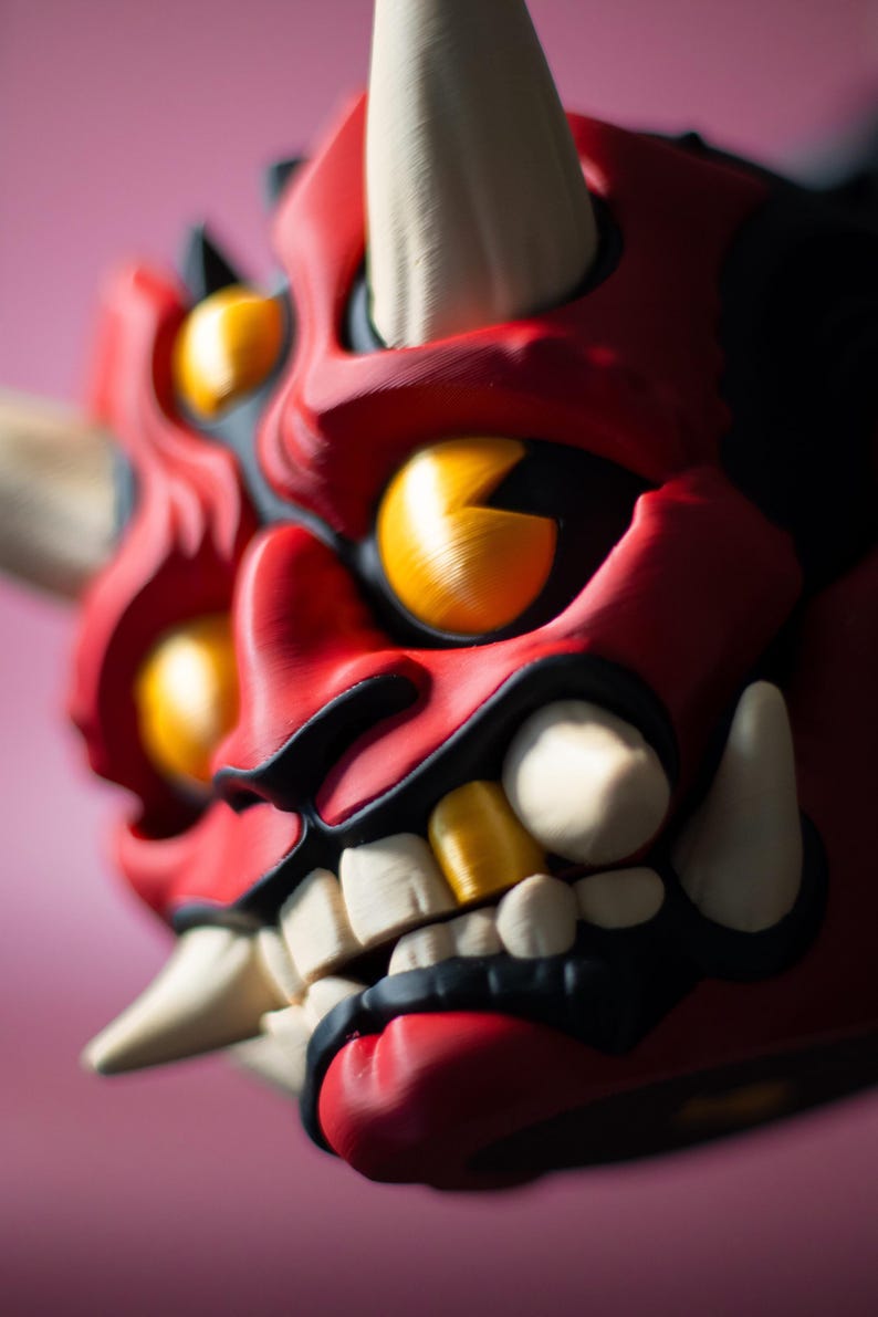 May include: A close-up of a red, black, and white Oni mask with yellow eyes and teeth. The mask has a fierce expression with sharp teeth and horns. The background is a soft pink.