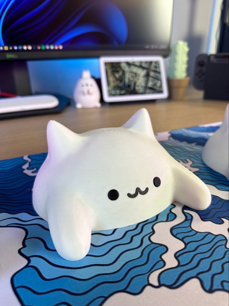 May include: A white, 3D-printed cat-shaped desk accessory with a smiling face. The cat has two pointed ears and outstretched arms. It rests on a blue and white wave-patterned mousepad. Other desk items are visible in the background.