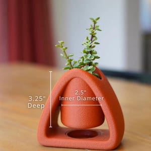 May include: A terracotta-colored, geometric planter with a small succulent plant. The planter has a 2.5-inch inner diameter and is 3.25 inches deep. The design features a suspended inner pot within a triangular frame.
