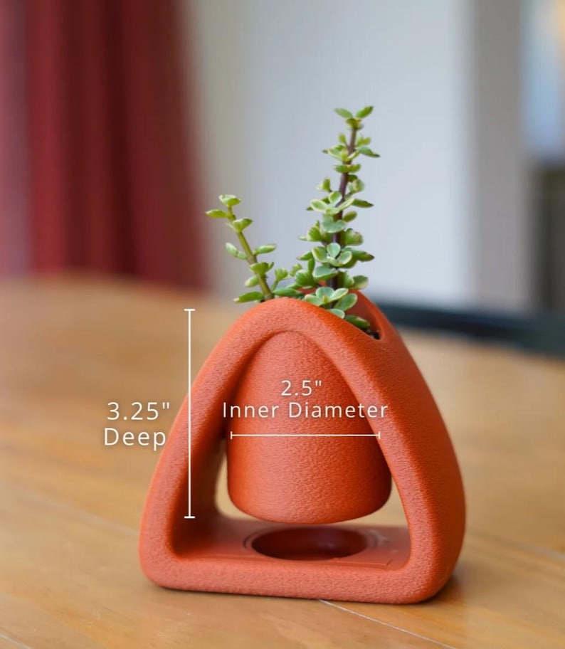 May include: A terracotta-colored, geometric planter with a small succulent plant. The planter has a 2.5-inch inner diameter and is 3.25 inches deep. The design features a suspended inner pot within a triangular frame.