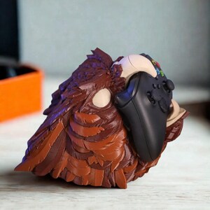 May include: A brown and tan sculpted owl-shaped controller holder. The holder is designed to cradle a black video game controller. The owl has detailed feather textures and a neutral color palette.