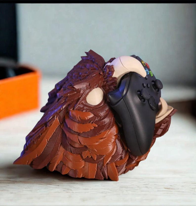 May include: A brown and tan sculpted owl-shaped controller holder. The holder is designed to cradle a black video game controller. The owl has detailed feather textures and a neutral color palette.