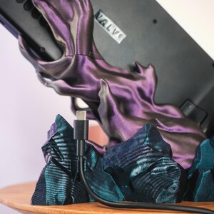 May include: A black gaming device with the word "VALVI" printed on it is resting on a purple and blue 3D printed stand. A black charging cable is plugged into the device.