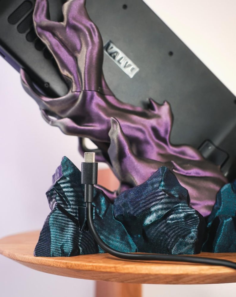 May include: A black gaming device with the word "VALVI" printed on it is resting on a purple and blue 3D printed stand. A black charging cable is plugged into the device.