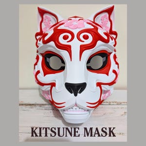 May include: A white Kitsune mask with red and pink floral accents. The mask features a black nose and eye openings, with the text "KITSUNE MASK" at the bottom. The mask is designed with a fox-like appearance.