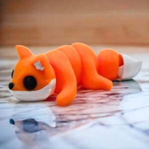 May include: A small orange and white 3D printed fox figurine lying on its side. The fox has large black eyes and a white muzzle.