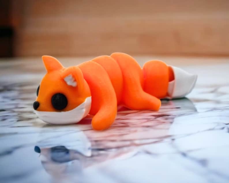 May include: A small orange and white 3D printed fox figurine lying on its side. The fox has large black eyes and a white muzzle.