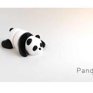 May include: A black and white panda figurine made of clay, lying on its belly with its head turned to the side. The panda has black ears, eyes, and paws. The figurine is on a white background. Panda Baby.
