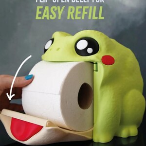 May include: A lime green frog-shaped toilet paper holder with large black and white eyes and a red cheek. The holder has a flip-open belly for easy refill, with the text "FLIP-OPEN BELLY FOR EASY REFILL" in green.