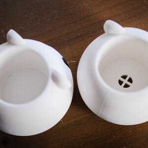 May include: Two white ceramic planters shaped like animal heads. The planter on the left has a black spot on the side. The planter on the right has a small drainage hole in the bottom.