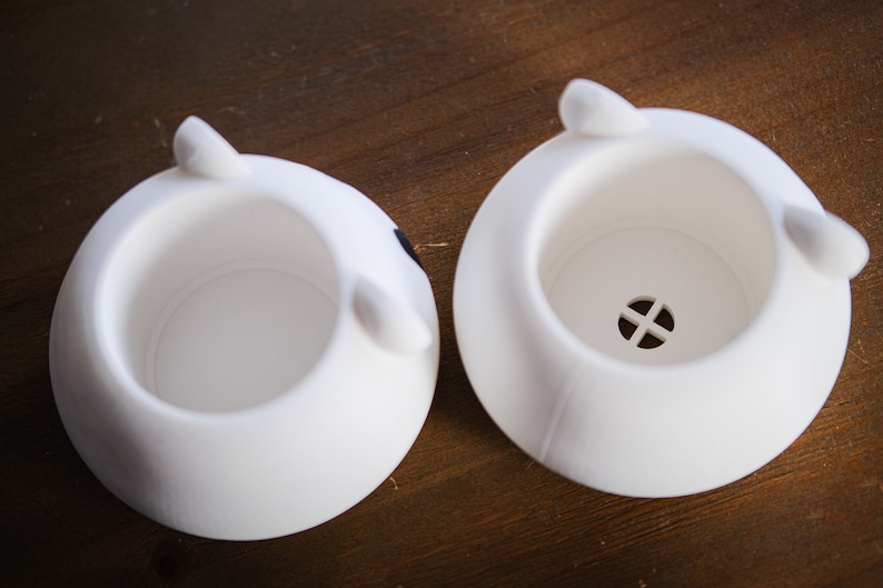 May include: Two white ceramic planters shaped like animal heads. The planter on the left has a black spot on the side. The planter on the right has a small drainage hole in the bottom.