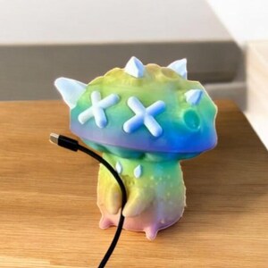 May include: A rainbow-colored 3D printed mushroom-shaped figurine with white X's on the top and a black charging cable plugged into the bottom.