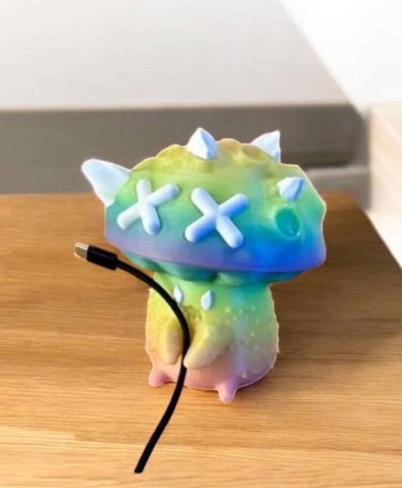 May include: A rainbow-colored 3D printed mushroom-shaped figurine with white X's on the top and a black charging cable plugged into the bottom.