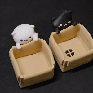 May include: Two small cardboard boxes with cat figurines peeking out of the top. The boxes are beige and have a small hole in the front. The cat figurines are white and black.