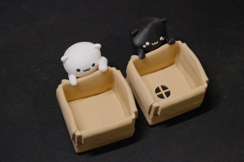 May include: Two small cardboard boxes with cat figurines peeking out of the top. The boxes are beige and have a small hole in the front. The cat figurines are white and black.