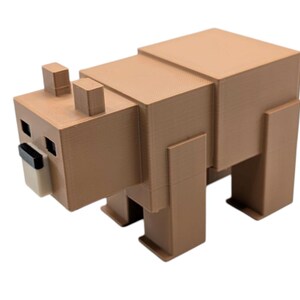 May include: A brown, blocky bear figurine with a geometric design. The bear has square eyes, a black nose, and a light tan snout. The ears are small, rectangular blocks. The bear is standing on four rectangular legs.
