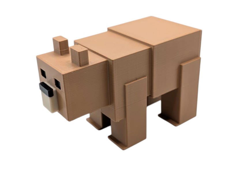 May include: A brown, blocky bear figurine with a geometric design. The bear has square eyes, a black nose, and a light tan snout. The ears are small, rectangular blocks. The bear is standing on four rectangular legs.