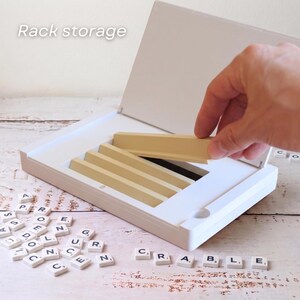 Puede incluir: Una caja blanca de juego de Scrabble con estantes de azulejos beige. El texto "Rack storage" es visible. Las fichas de letras individuales est&aacute;n esparcidas alrededor de la caja, con la palabra "SCRABBLE" escrita. Una mano est&aacute; colocando un estante de fichas.