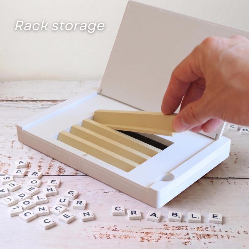 Puede incluir: Una caja blanca de juego de Scrabble con estantes de azulejos beige. El texto "Rack storage" es visible. Las fichas de letras individuales est&aacute;n esparcidas alrededor de la caja, con la palabra "SCRABBLE" escrita. Una mano est&aacute; colocando un estante de fichas.