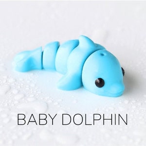 May include: A light blue plastic baby dolphin toy with black eyes. The dolphin is on a white surface with water droplets.