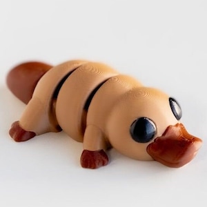Puede incluir: Una figura de ornitorrinco impresa en 3D de color marr&oacute;n y beige con ojos negros. El ornitorrinco est&aacute; acostado boca abajo con la cabeza girada hacia la derecha.