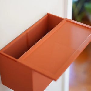 May include: An open, rectangular, orange-colored wall-mounted storage box with a lid. The box is divided into two compartments. The lid is partially open, revealing the interior. The box is mounted on a white wall.