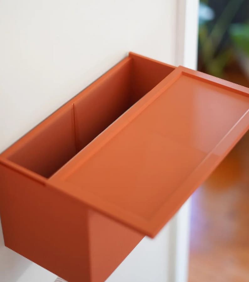 May include: An open, rectangular, orange-colored wall-mounted storage box with a lid. The box is divided into two compartments. The lid is partially open, revealing the interior. The box is mounted on a white wall.