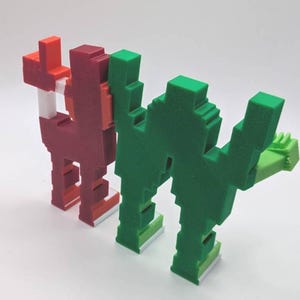May include: Two pixelated sculptures, one red and one green, stand side-by-side. The red sculpture has a white accent and the green one has a light green hand. Both have a blocky, geometric design, reminiscent of retro video game characters.