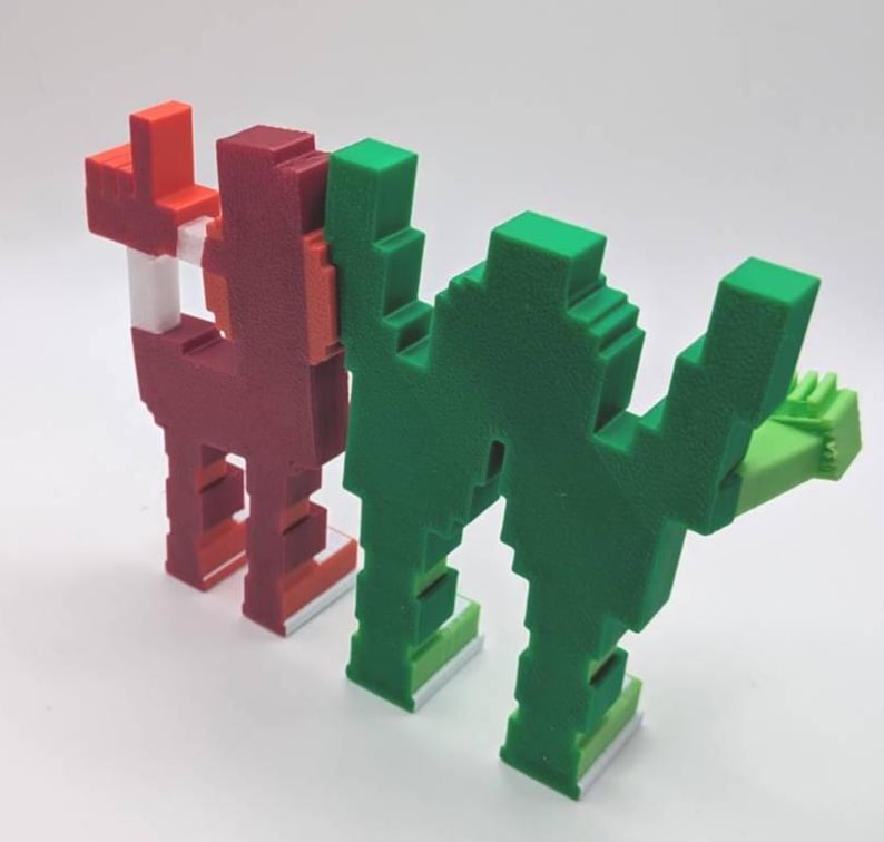 May include: Two pixelated sculptures, one red and one green, stand side-by-side. The red sculpture has a white accent and the green one has a light green hand. Both have a blocky, geometric design, reminiscent of retro video game characters.