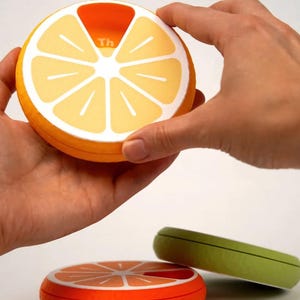 May include: A round orange pill organizer shaped like a citrus fruit slice, held by hands. The top section is open, revealing a compartment labeled "Th". Two other pill organizers, one orange and one green, are also visible.