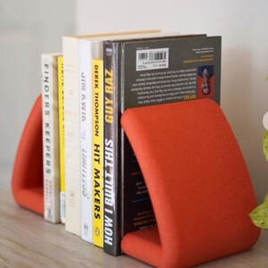 May include: A set of books held upright by two orange bookends. The books have various titles, including "HOW I BUILT THIS" and "FINDERS KEEPERS". The bookends are a solid, curved design, providing a modern aesthetic to the bookshelf.