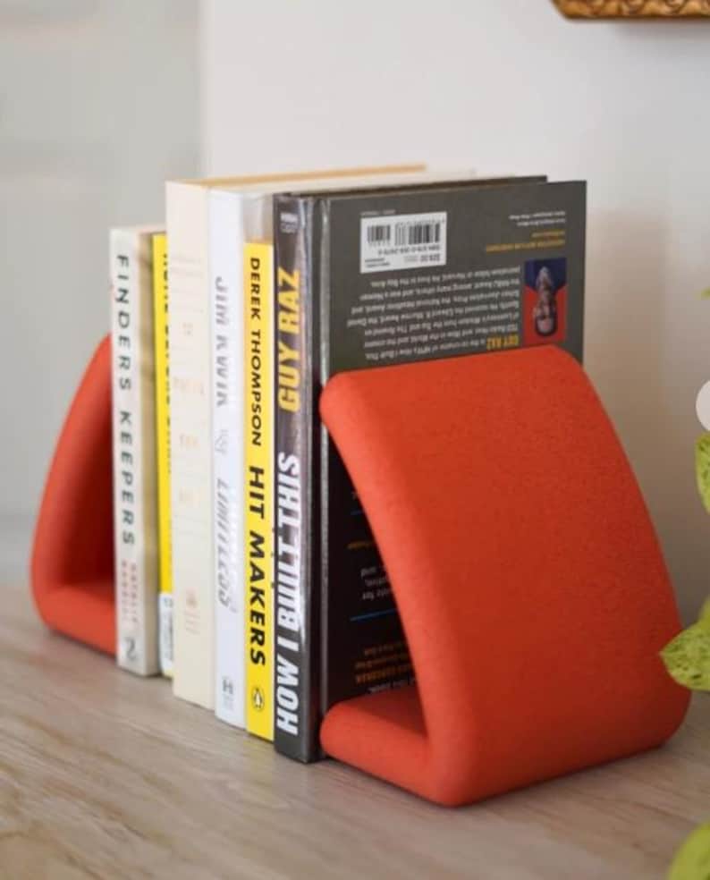 May include: A set of books held upright by two orange bookends. The books have various titles, including "HOW I BUILT THIS" and "FINDERS KEEPERS". The bookends are a solid, curved design, providing a modern aesthetic to the bookshelf.
