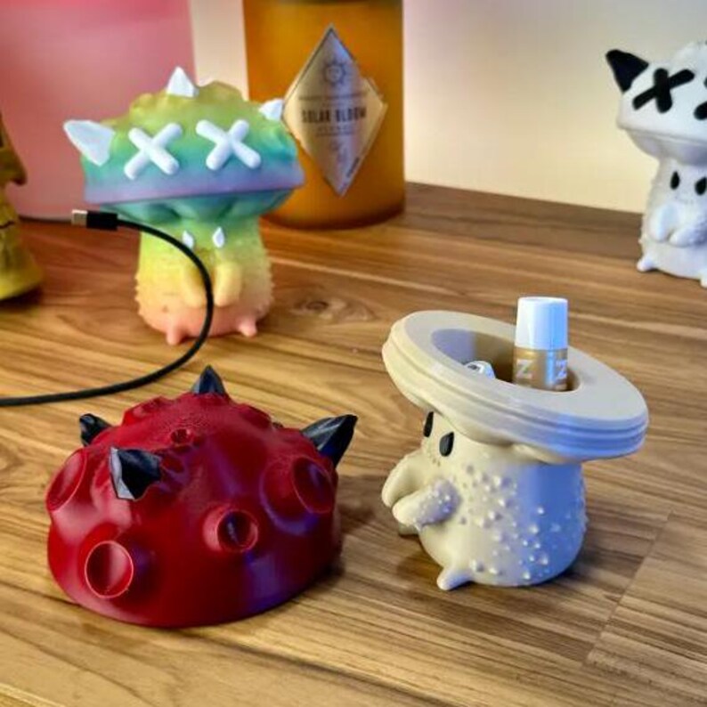 May include: Four colorful 3D printed figurines, each with a unique design. The figurines are made of a smooth, plastic-like material. The figurine on the left is red with black accents and resembles a planet. The figurine in the center is beige with a wide, flat top and resembles a mushroom. The figurine on the right is white with black accents and resembles a creature with a large head. The figurine in the back is rainbow colored with white accents and resembles a mushroom.