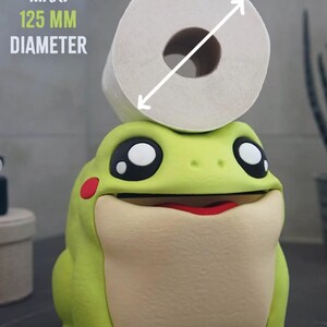 May include: A whimsical, lime green frog-shaped toilet paper holder with a roll of white toilet paper on top. The frog has large black eyes, a red mouth, and a maximum diameter of 125 mm. The holder adds a fun touch to bathroom decor.