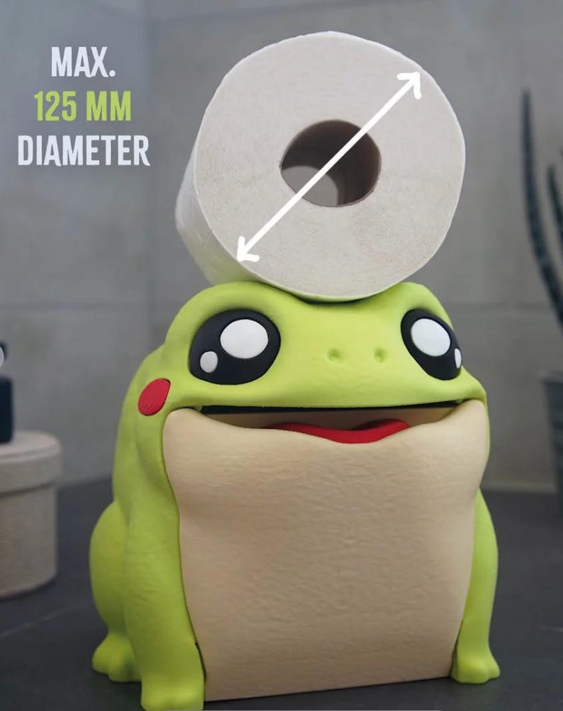 May include: A whimsical, lime green frog-shaped toilet paper holder with a roll of white toilet paper on top. The frog has large black eyes, a red mouth, and a maximum diameter of 125 mm. The holder adds a fun touch to bathroom decor.